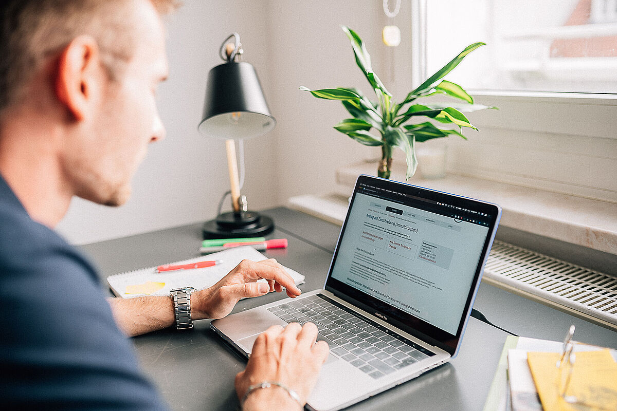 Ein Mann sitzt an einem Laptop an einem Schreibtisch. Im Hintergrund sieht man eine Zimmerpflanze und eine Lampe. 