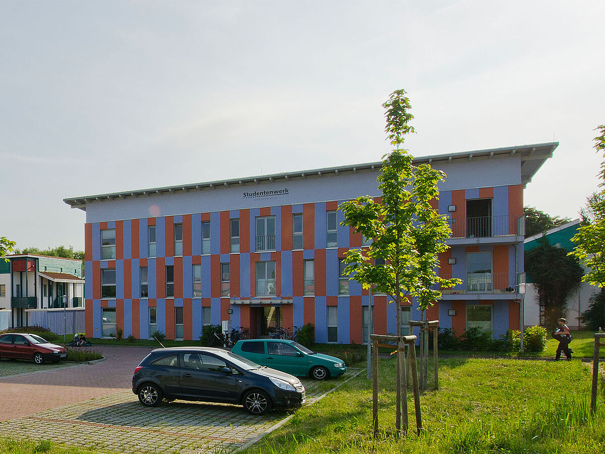 Modernes, rechteckiges Flachdachgebäude des Studentenwohnheim mit Parkplatz und Begrünung im Vordergrund.