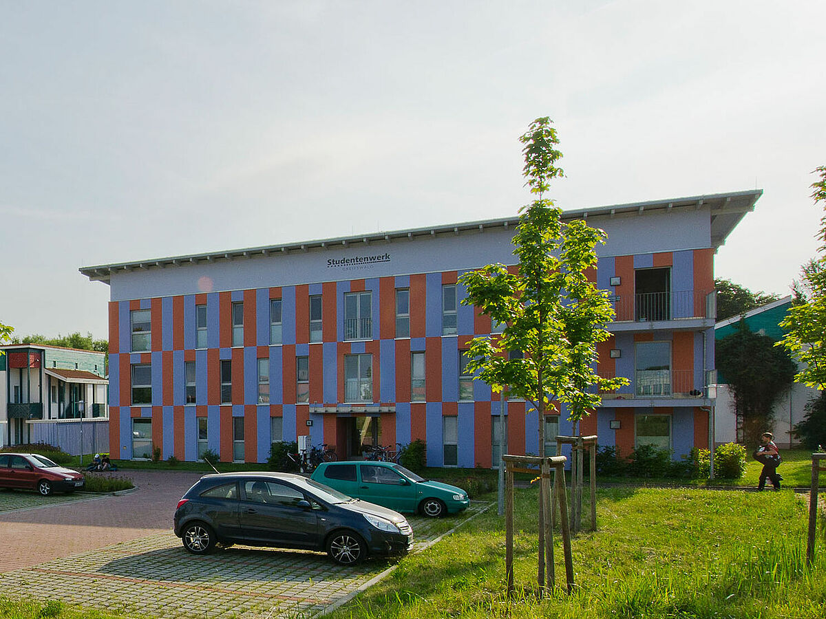 Modernes, rechteckiges Flachdachgebäude des Studentenwohnheim mit Parkplatz und Begrünung im Vordergrund.