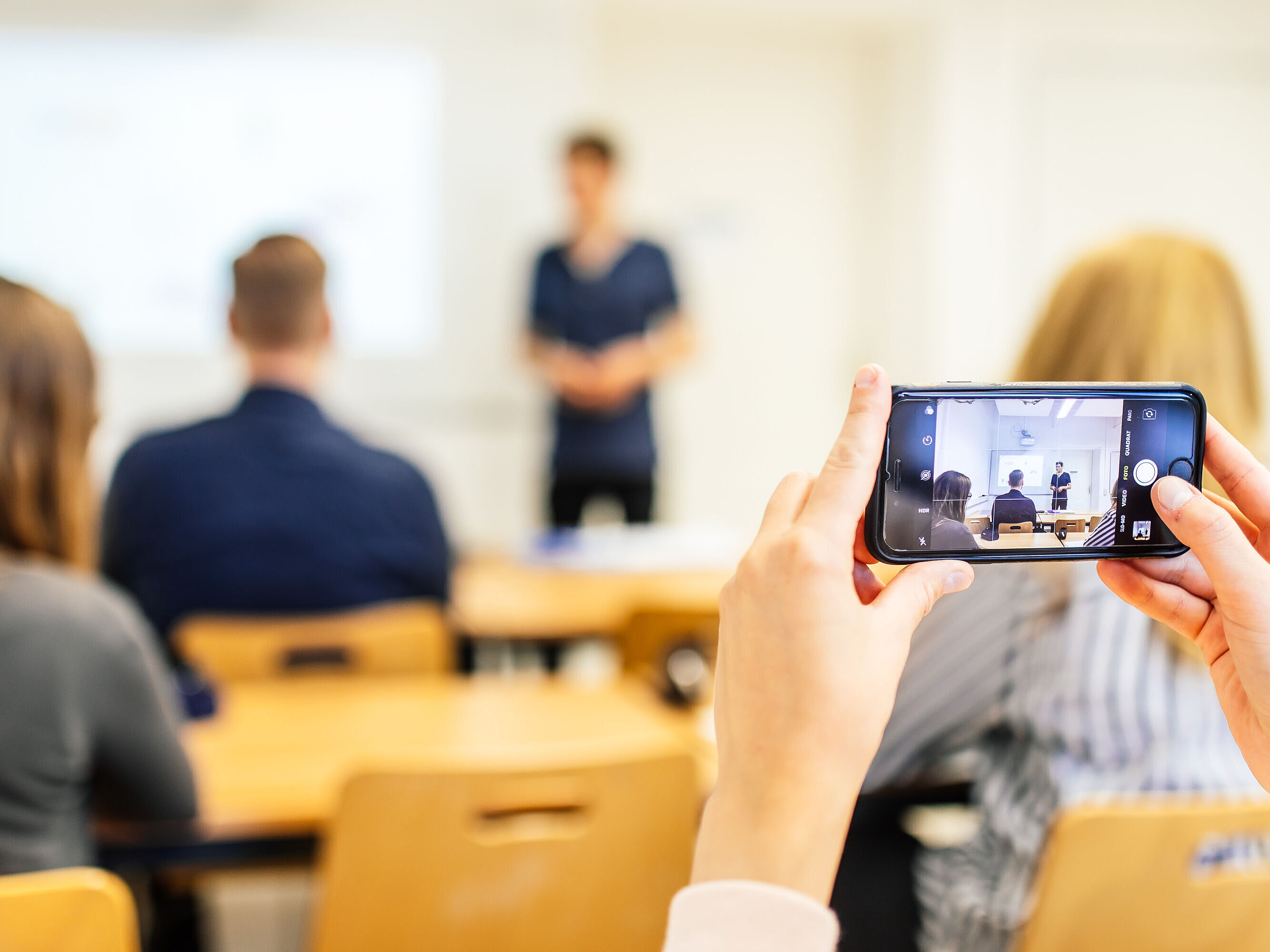 Symbolbild Studium Im Vordergrund hält eine Person ein Smartphone in der Hand, im Hintergrund sind weitere Personen sehr verschwommen zu sehen.