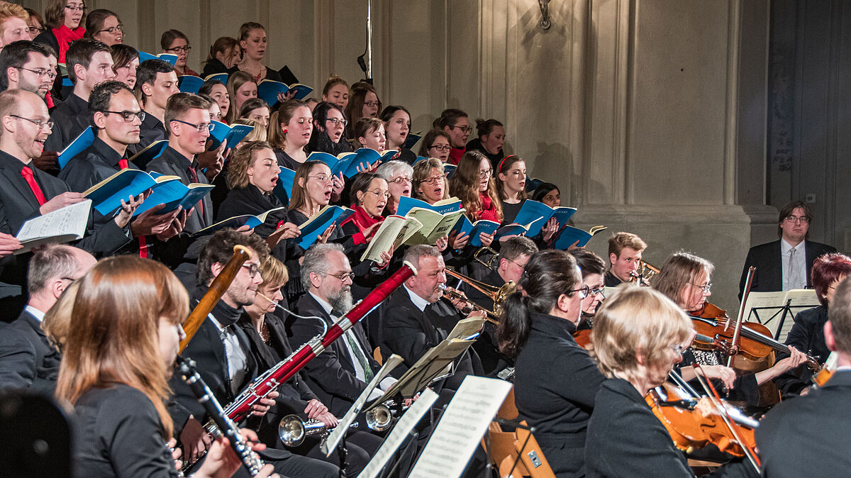 Eine Vielzahl von Menschen mit Instrumenten und Notenständern in schwarzer Kleidung auf der Bühne.