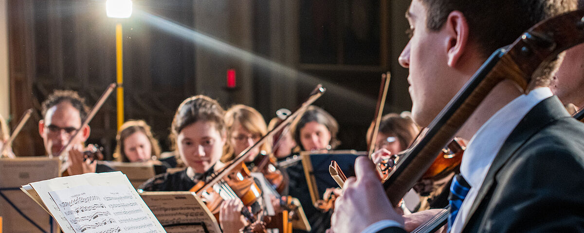 Vorne rechts sitzt in Nahaufnahme ein Mann mit aufgeschlagenen Notenblättern, der Cello spielt. im Hintergrund sieht man weitere Musizierende des Universitätssinfonieorchesters.