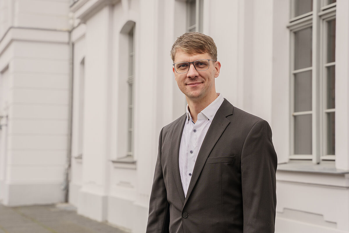 Porträt Prof. Hendrik Lohse-Bossenz Ein Mannn steht vor einem weißen Gebäude und lächelt in die Kamera.