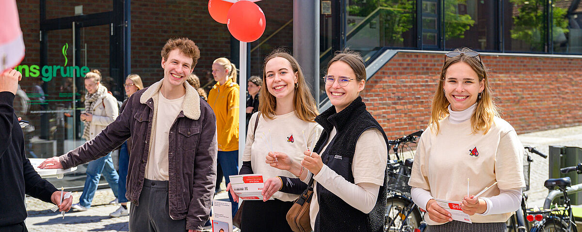 Four students are standing under a white tent in front of the “ins grüne” cafeteria on the Loefflerstraße campus. They are holding information material from the DKMs in their hands and smiling at the camera.
