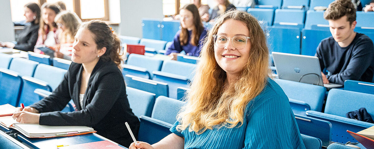 Im Hörsaal sitzen viele Studierende. Im Vordergrund sitzt eine blonde Frau in einem blauen Kleid.