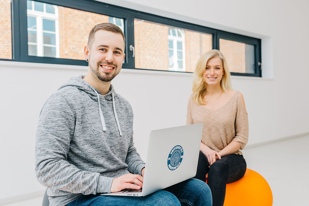 Eine Studentin und ein Student sitzen im Hörsaalgebäude. Er hat ein Notebook auf dem Schoß.