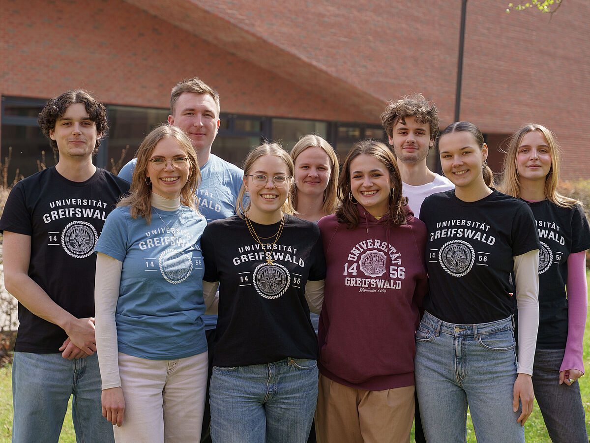 Gruppenbild der Campusspezialist*innen 2026 auf dem Campus Loefflerstraße. Abgebildet sind neun Personen vor einem großen Hörsaalgebäude aus Backstein. Vorne stehen vier Campusspezialistinnen. In der Reihe dahinter stehen drei männliche und zwei weibliche Campusspezialist*innen. Sie tragen T-Shirts der Universität in verschiedenen Farben und lächeln freundlich in die Kamera. 