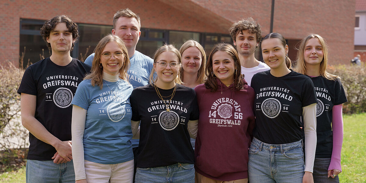 Gruppenbild der Campusspezialist*innen 2026 auf dem Campus Loefflerstraße. Abgebildet sind neun Personen vor einem großen Hörsaalgebäude aus Backstein. Vorne stehen vier Campusspezialistinnen. In der Reihe dahinter stehen drei männliche und zwei weibliche Campusspezialist*innen. Sie tragen T-Shirts der Universität in verschiedenen Farben und lächeln freundlich in die Kamera. 