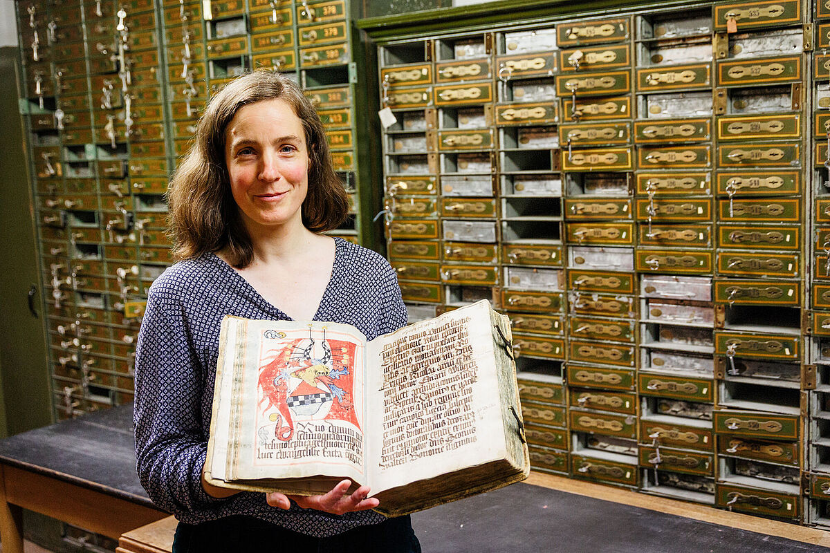 Dr. Elisabeth Heigl steht vor einem alten Archivschrank und hält ein historisches Buch geöffnet in den Händen.