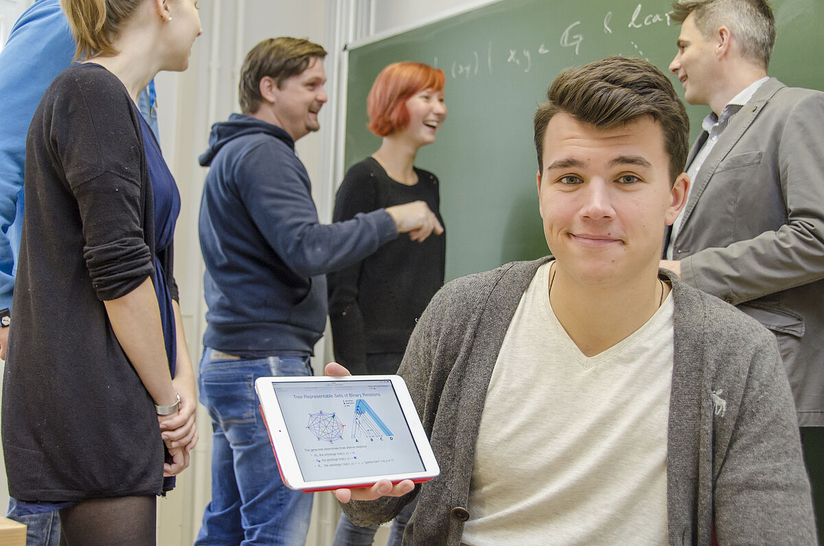 Symbolbild Mathematik Studierende mit Dozierender Person vor einer grüner Tafel im Vordergrund hält ein junger Mann ein Tablet in der Hand