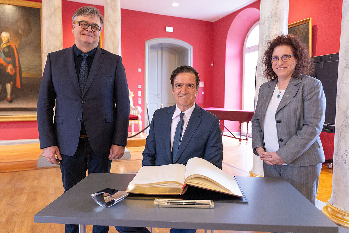 The image shows Prof. Dr. Marko Pantermöller, H.E. Ambassador Dr. Kai Sauer, and Rector Prof. Dr. Katharina Riedel during the signing of the guest book in the university's Aula