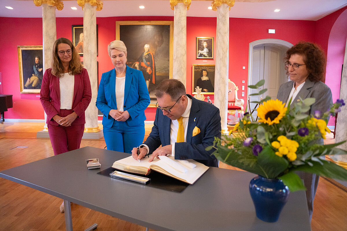 The picture was taken in the university's historic Aula. It shows (from left) Minister of Science Bettina Martin, the Ukrainian Ambassador H.E. Oleksii Makeiev, Minister-President Manuela Schwesig and Rector Prof. Dr. Katharina Riedel. The Ukrainian Ambassador is making an entry into the university's guest book.