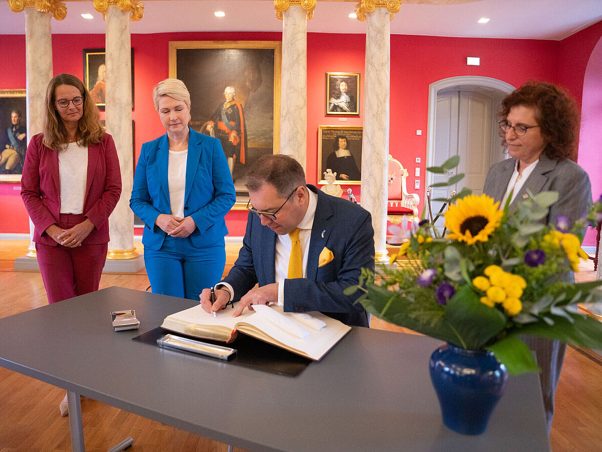 Das Bild ist in der Historischen Aula der Universität aufgenommen. Zu sehen sind von links nach rechts die Wissenschaftsministerin Bettina Martin, der Ukrainische Botschafter S.E. Oleksii Makeiev, die Ministerpräsidentin Manuela Schwesig und die Rektorin Prof. Dr. Katharina Riedel.n.r.: Auf dem Bild sitzt der ukrainische Botschafter am Tisch und trägt sich in das Ehrenbuch der Universität ein. Links neben ihm stehen die Ministerpräsidentin Manuela Schwesig und die Wissenschaftsministerin Bettina Martin. Rechts von ihm steht Rektorin Prof. Dr. Katharina Riedel.