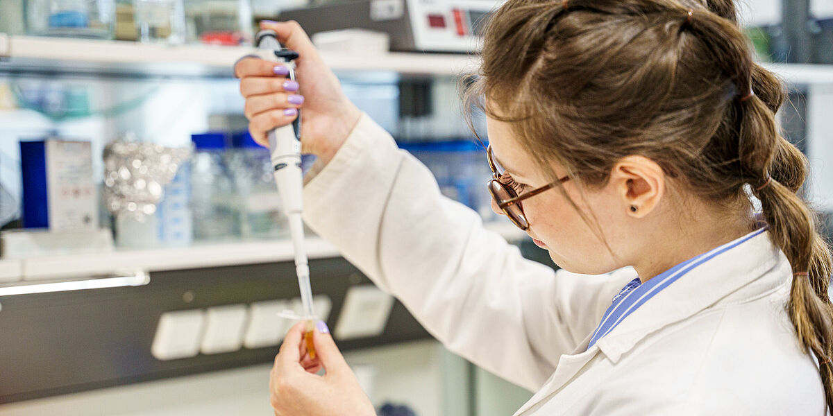 Fun Gene Labor 2026 Eine Studierende im einem Laborkittel hält eine Pipette hoch und tropft etwas in einen kleinen Auffangbehälter.