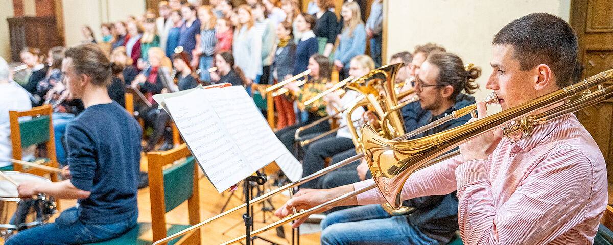 Im Vordergrund sitzen Männer mit Blasinstrumenten, im Hintergrund steht der Chor.