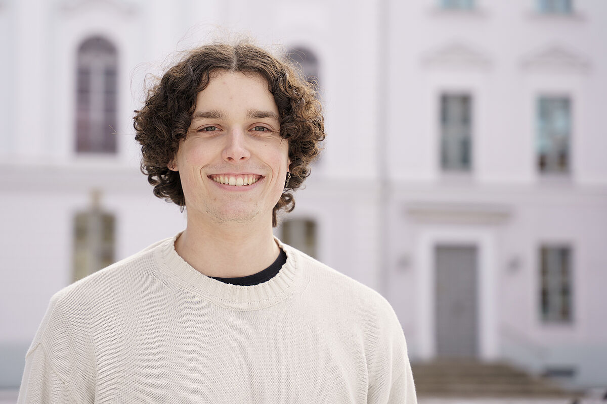 Ein Poträt von Eric Engelhardt, der in die Kamera lächelt und vor dem Universitätshauptgebäude steht.