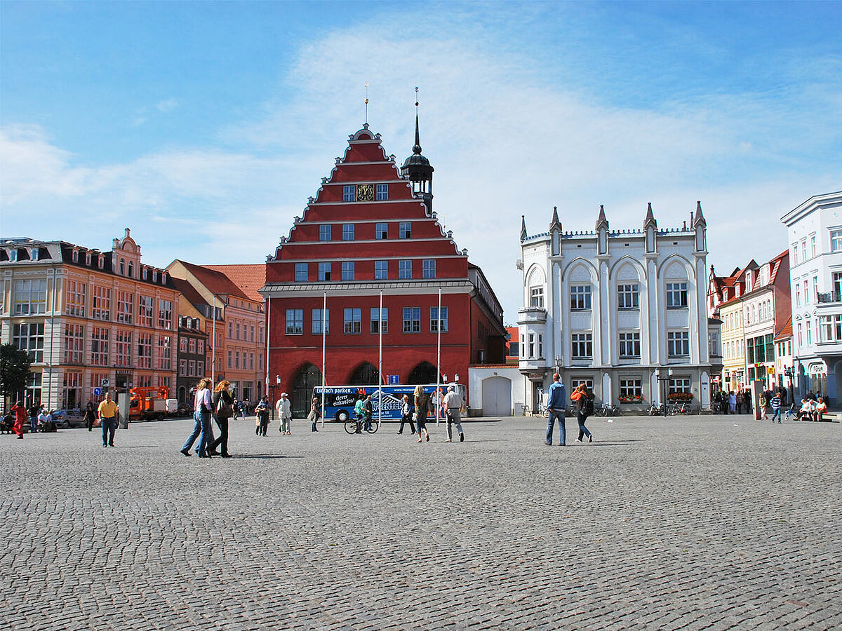 Blick über den Greifswalder Markt auf das Rathaus der Stadt. Es ist schönes Wetter und Leute gehen über den Markt.