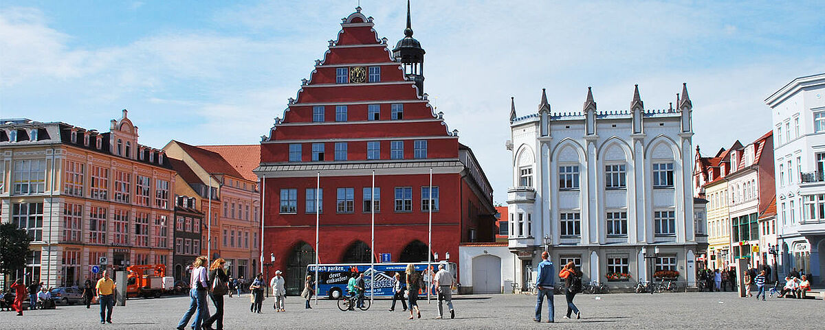 Blick über den Greifswalder Markt auf das Rathaus der Stadt. Es ist schönes Wetter und Leute gehen über den Markt.