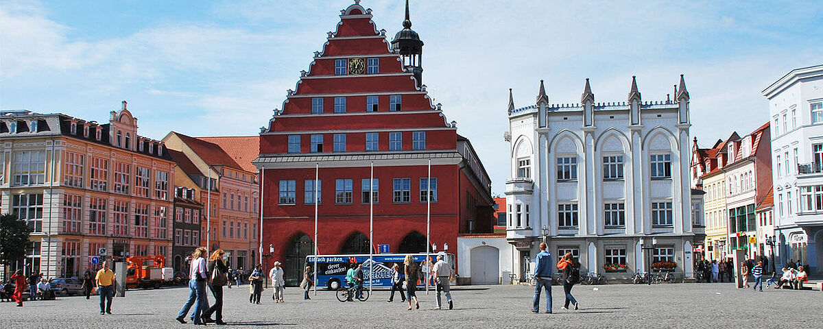 Blick über den Greifswalder Markt auf das Rathaus der Stadt. Es ist schönes Wetter und Leute gehen über den Markt.