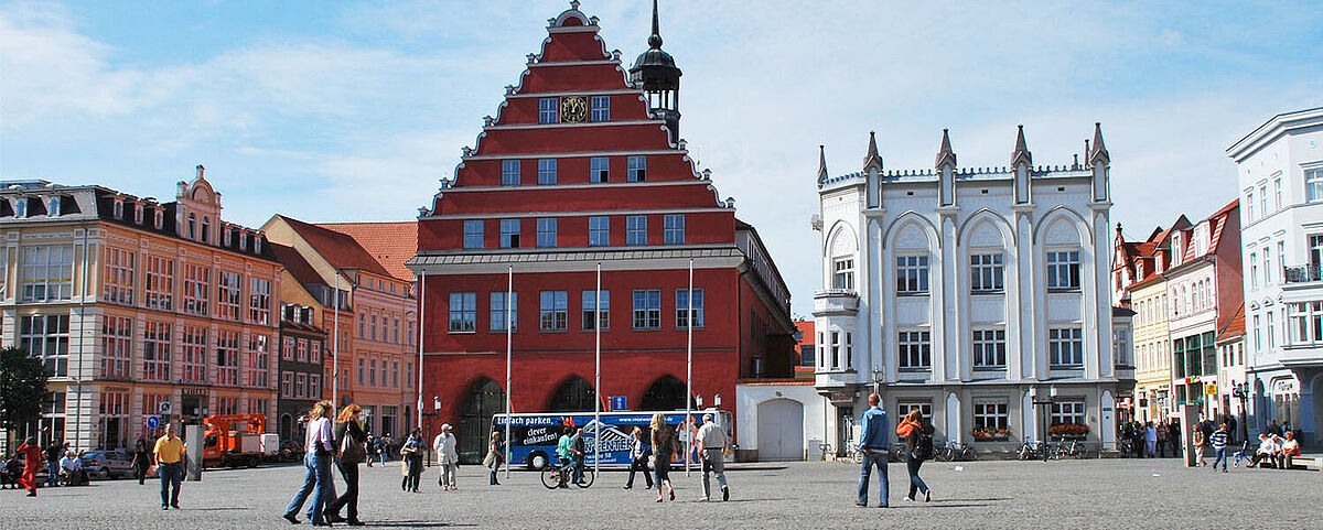 Blick über den Greifswalder Markt auf das Rathaus der Stadt. Es ist schönes Wetter und Leute gehen über den Markt.