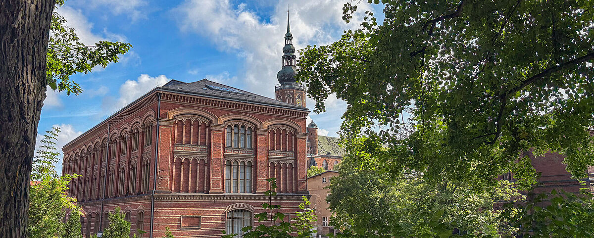Zwischen einem Rahmen aus grünen Blättern steht die rote, backsteinerne Alte Universitätsbibliothek.