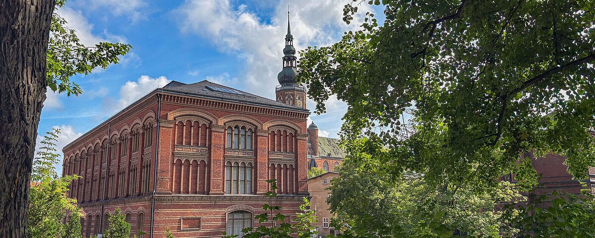 Zwischen einem Rahmen aus grünen Blättern steht die rote, backsteinerne Alte Universitätsbibliothek.