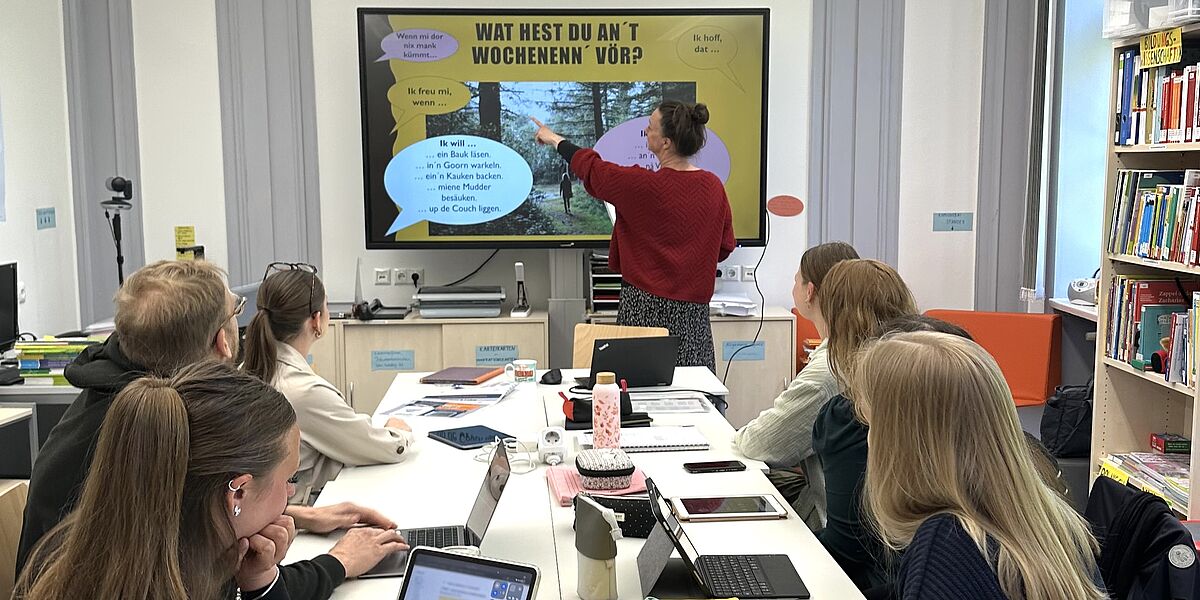 Unterricht im Grundschullehramt Niederdeutsch 2026 Wir sehen eine Unterrichtssituation. Studierende sitzen an einem großen Tisch und schauen in die Richtung der Tafel. An der Tafel wird Ihnen ein Schaubilld auf Niederdeutsch erklärt.