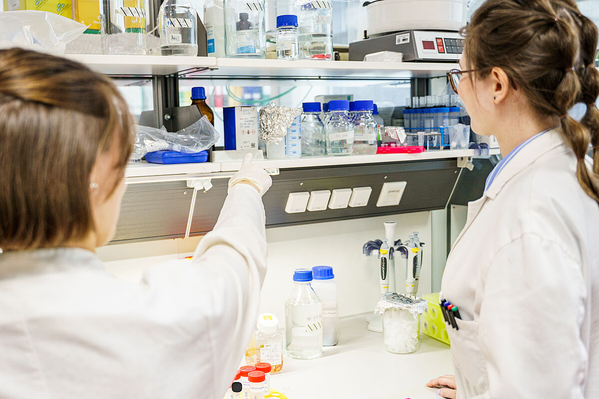 Das Bild zeigt zwei Forscherinnen, die in einem Laborraum mit verschiedenen Chemikalien und Instrumenten arbeiten.