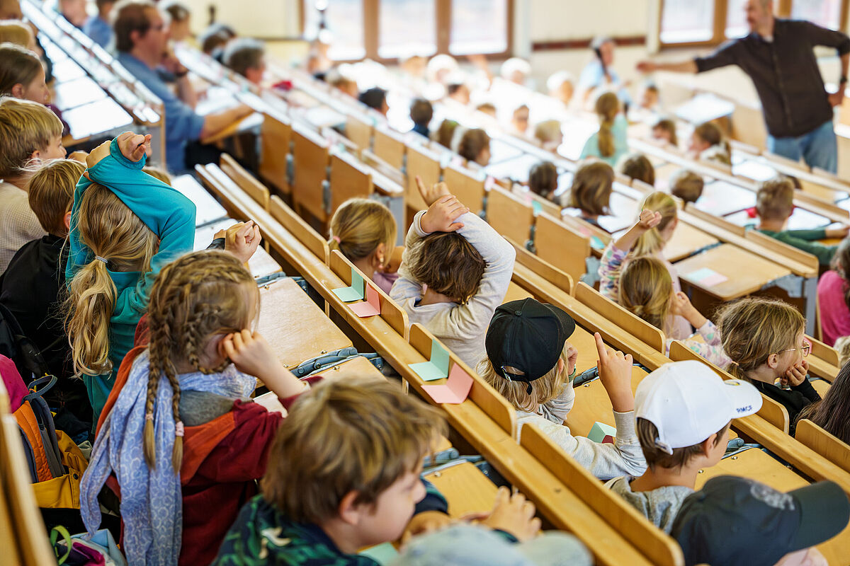 In einem hellen Hörsaal sitzen Schüler*innen, die einem Vortrag lauschen. Einige melden sich.