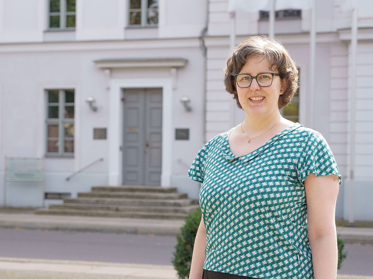 Frau Dr. Amrei Koch steht vor dem hellen Universitätshauptgebäude in Greifswald.