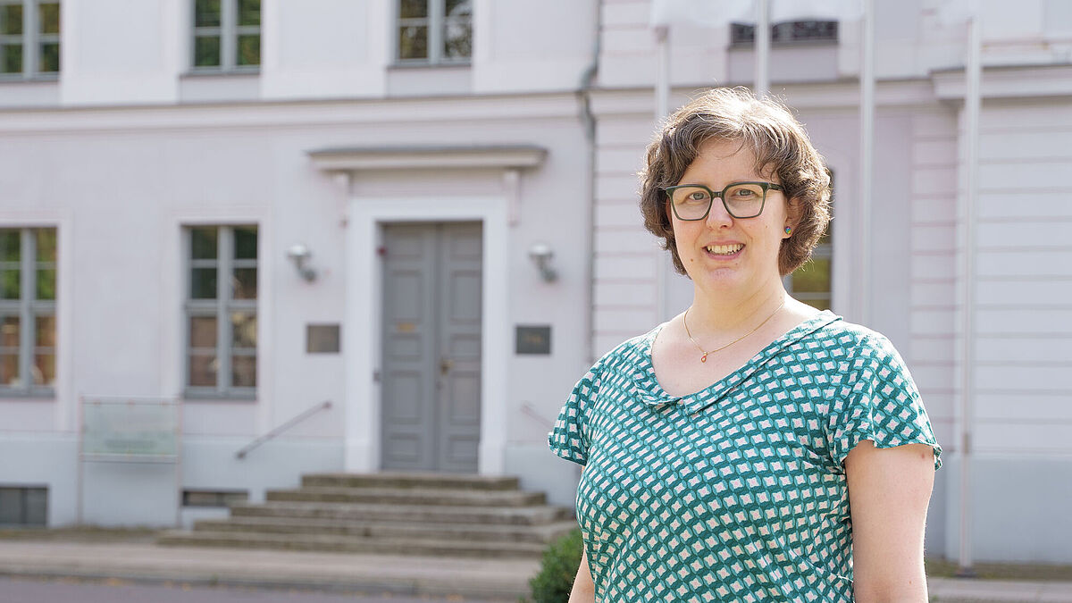 Porträt Dr. Amrei Koch Frau Dr. Amrei Koch steht vor dem hellen Universitätshauptgebäude in Greifswald.