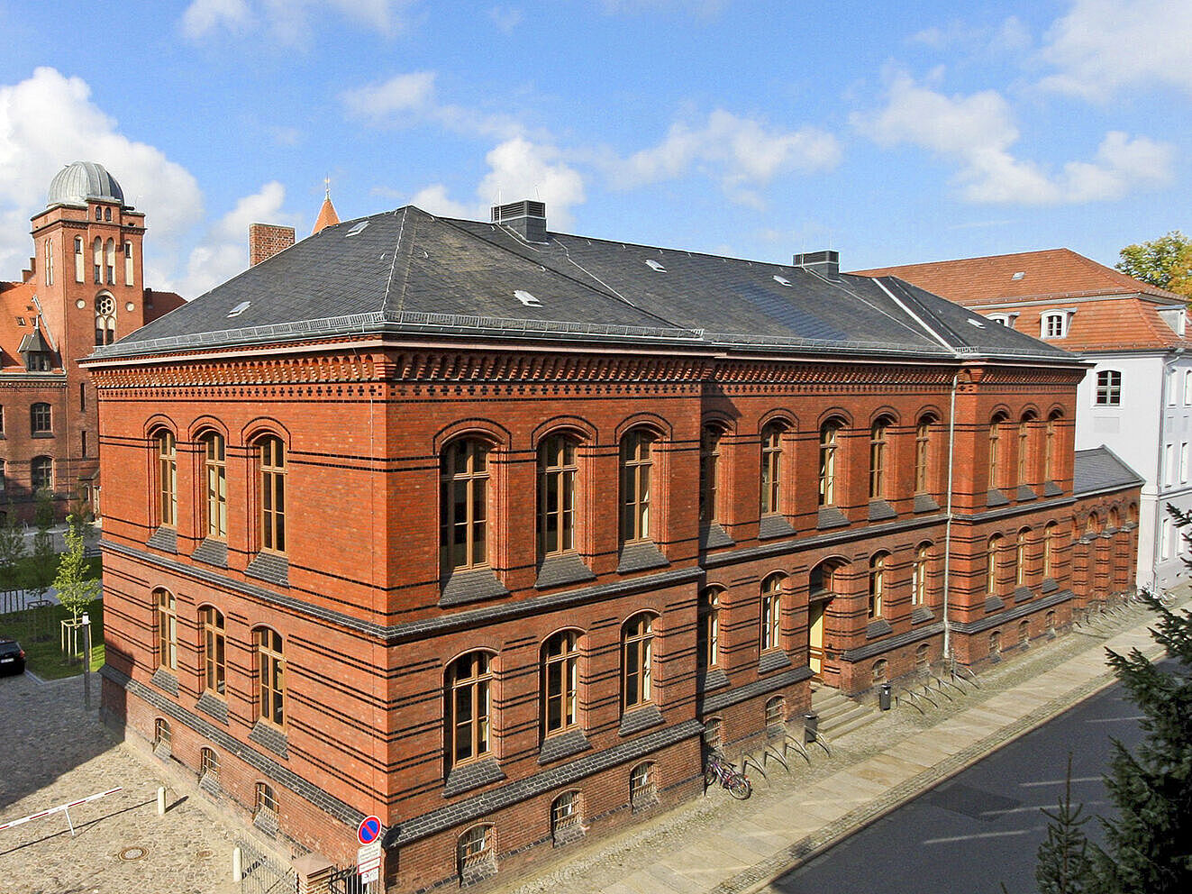 Symbolbild Altes Audimax Das Bild zeigt das Alte Audimax, ein historisches, zweistöckiges Backsteingebäude mit großen Fenstern und dunklem Schieferdach, umgeben von weiteren Gebäuden. Der Himmel ist blau mit vereinzelten weißen Wolken.