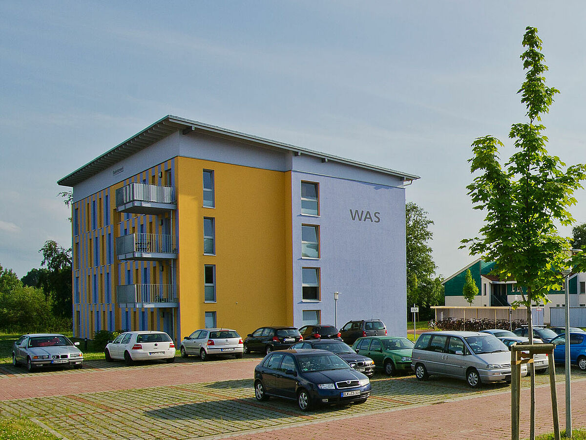 Wohnheim Fleischerwiese. Modernes, rechteckiges Flachdachgebäude des Studentenwohnheim mit Parkplatz im Vordergrund.