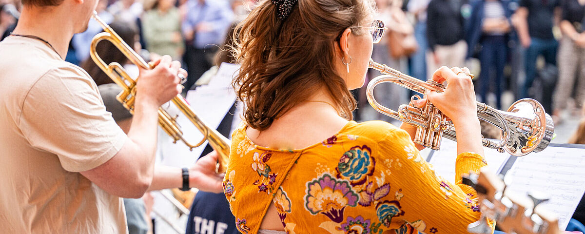 Ein Mann und eine Frau sind von hinten zu sehen. Sie stehen mit Blasinstrumenten auf der Bühne, im Hintergrund steht das Publikum.