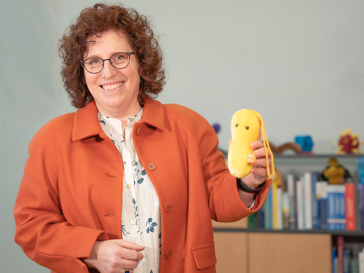 Auf dem Bild ist Prof. Dr. Katharina Riedel abgebildet. In der Hand hält sie ein Plüschtier, eine Mikrobe.