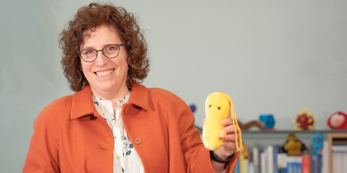 Auf dem Bild ist Prof. Dr. Katharina Riedel abgebildet. In der Hand hält sie ein Plüschtier, eine Mikrobe.