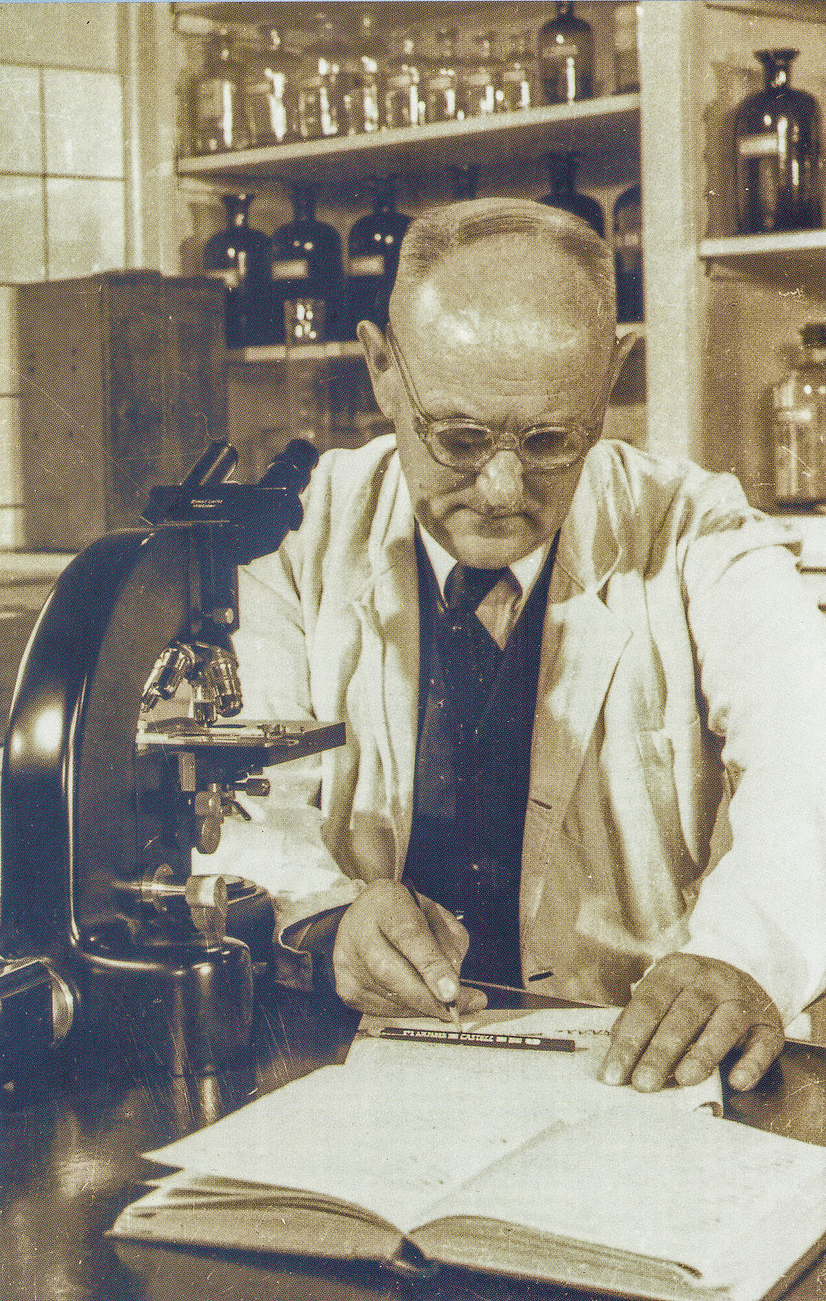 Ein Mann im Laborkittel beim Eintragen mikroskopischer Befunde in eine Laborjournal. Neben ihm steht links ein Mikroskop.