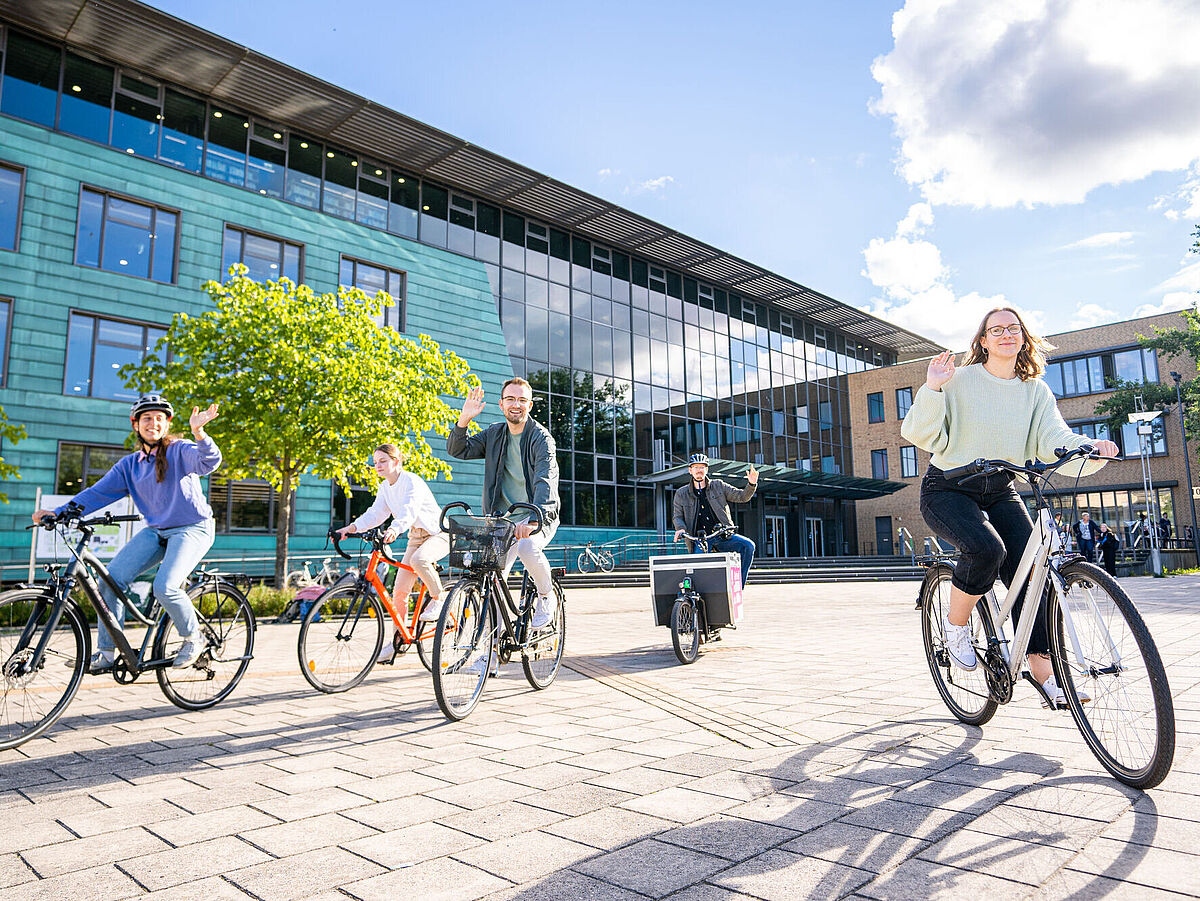 Studierende fahren mit dem Fahrrad und winken fröhlich in die Kamera. Im Hintergrund ist die Zentrale Universitätsbibliothek zu sehen.