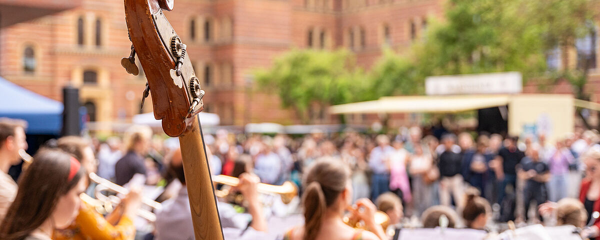Der Hals einer Violine ist im Vodergrund zu sehen. Im Hintergrund sitzen schemenhaft Menschen, die dem sommerlichen Konzert beiwohnen.