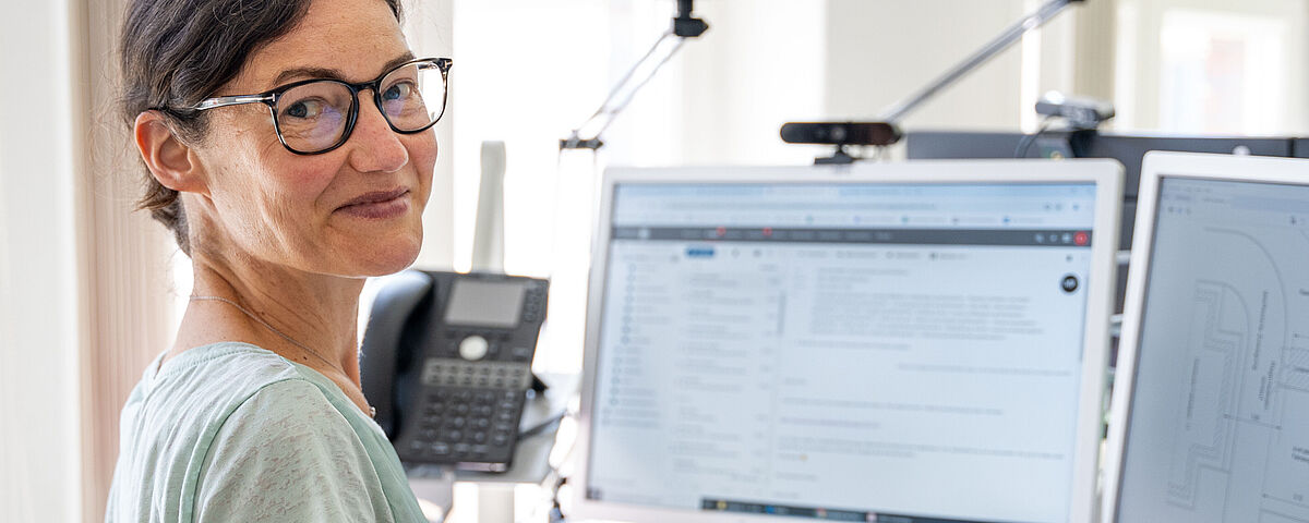Eine Frau mittleren Alters sitzt vor einem PC mit großem Monitor und lächelt freundlich in die Kamera.