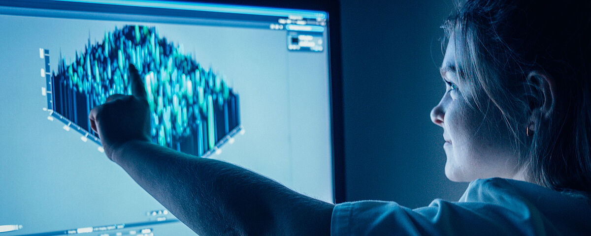 Eine Wissenschaftlerin steht vor einem Monitor mit einer 3-D-Grafik und zeigt auf einen Bildausschnitt.