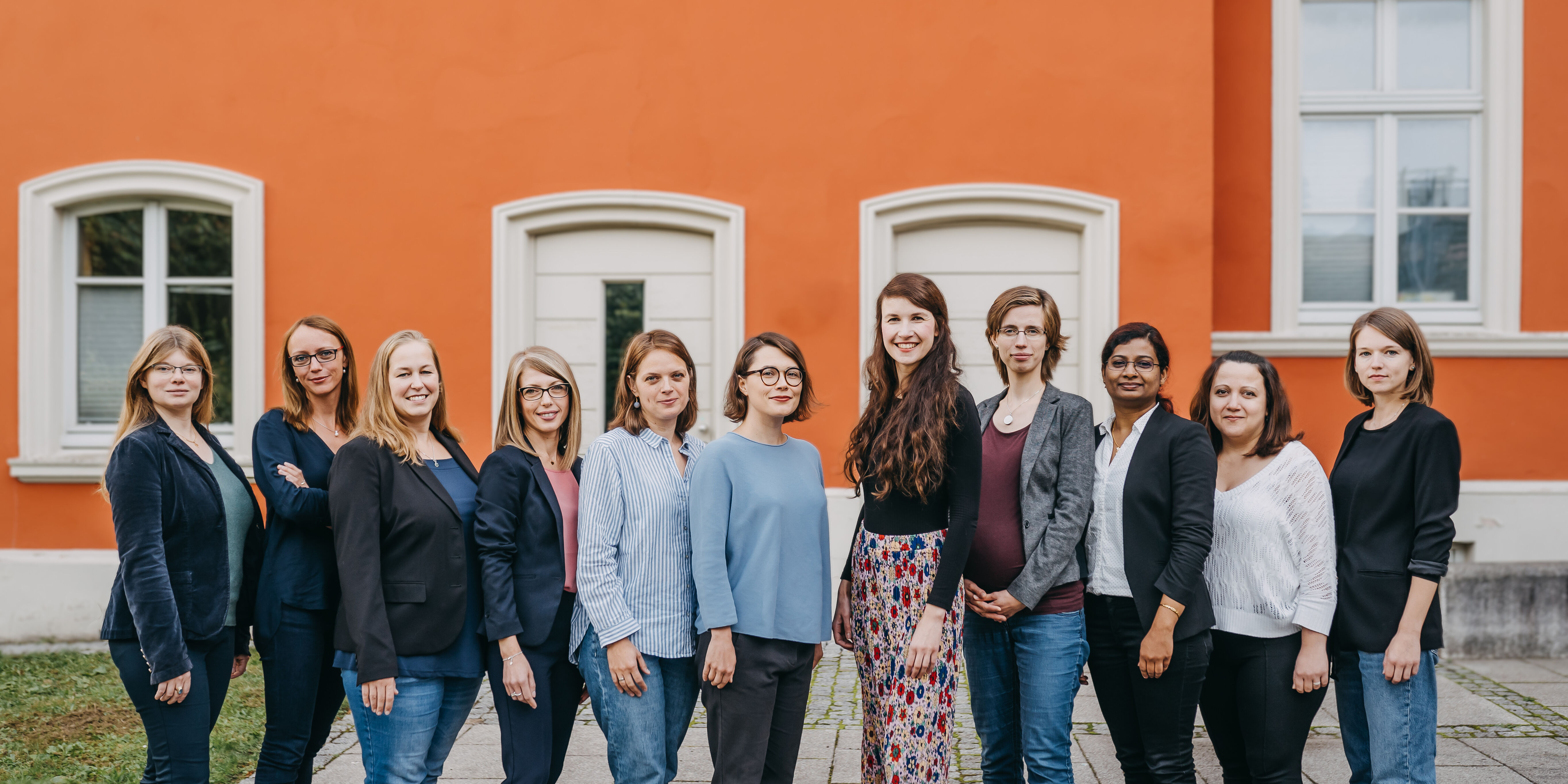 11 Frauen stehen nebeneinander, sie stehen vor einer roten Häuserwand, sie schauen in die Kamera