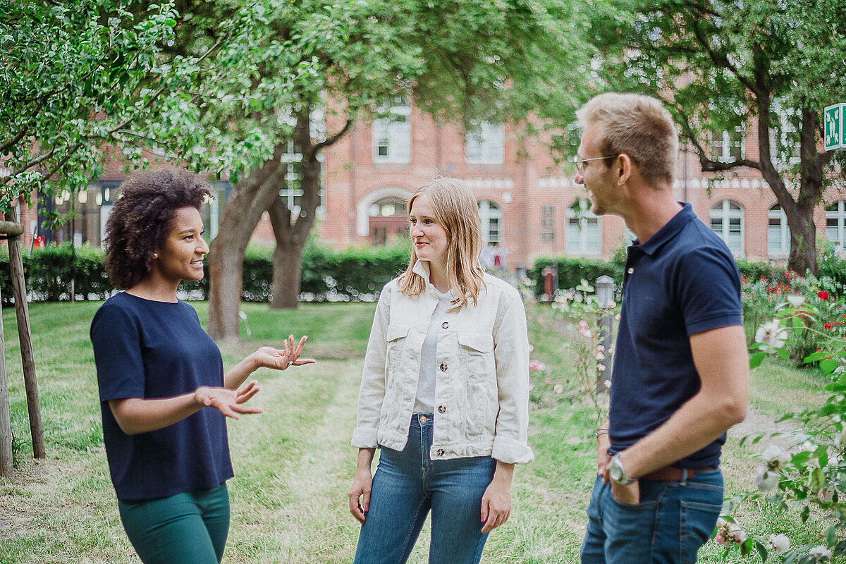 Studierende im Gespräch; Drei Studierende unterhalten sich am Campus Loefflerstraße angeregt miteinander. Sie stehen in einem kleinen Garten/Grünfläche, während man im Hintergrund die historischen Backsteinbauten erkennt.