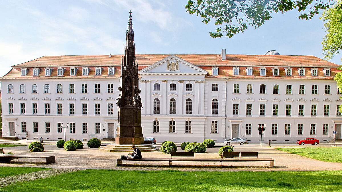 Vorderansicht des Universitätshauptgebäudes mit Rubenowdenkmal im Sommer bei grünem Gras, blauem Himmel und einem grünen Baum.