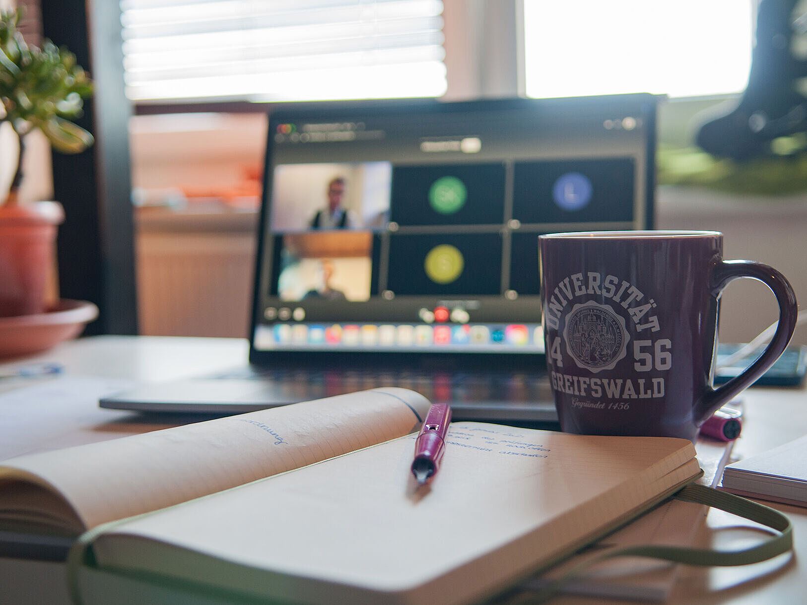 Ein Schreibtisch mit einem aufgeklappten Laptop, auf dem ein Videocall läuft, einer Tasse mit Universitätslogo und einem offenen Notizbuch mit Stift.