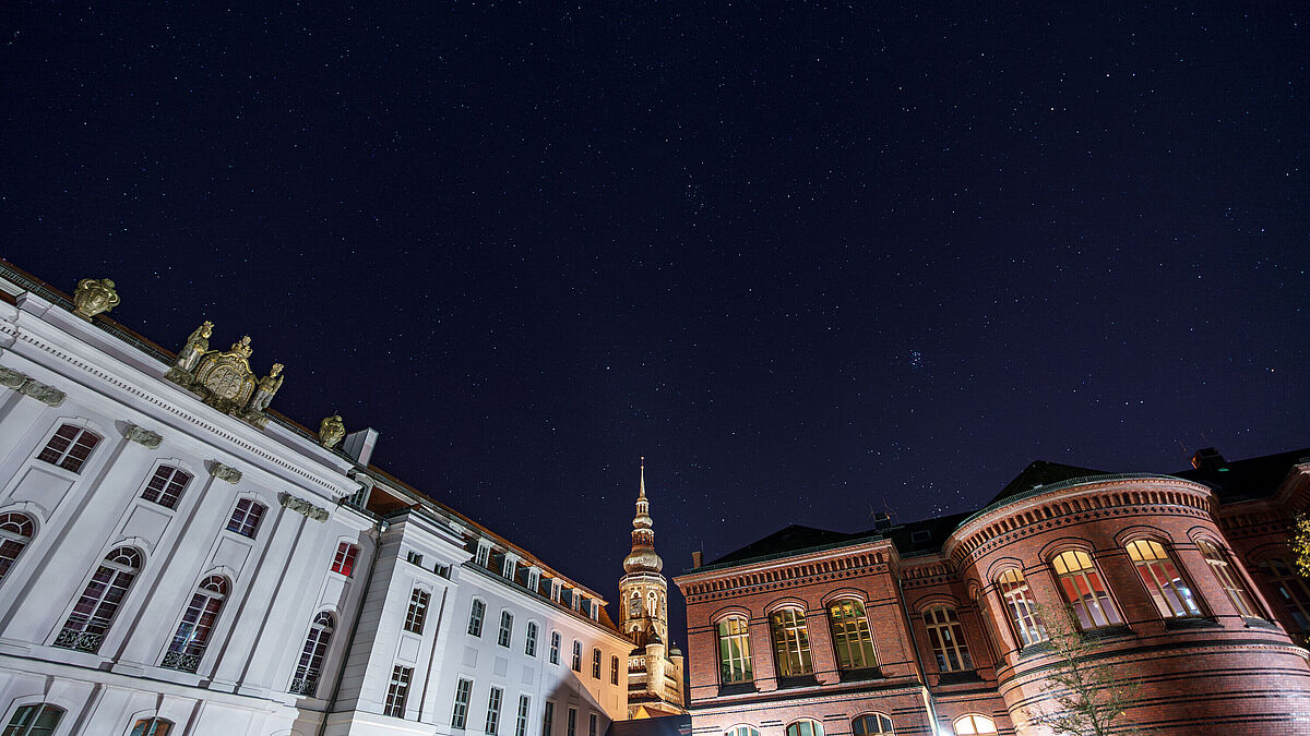 The image shows the Historic Campus with a starry sky. The Main Building is on the left and the Altes Audimax on the right. The St. Nikolai cathedral can be seen in the centre