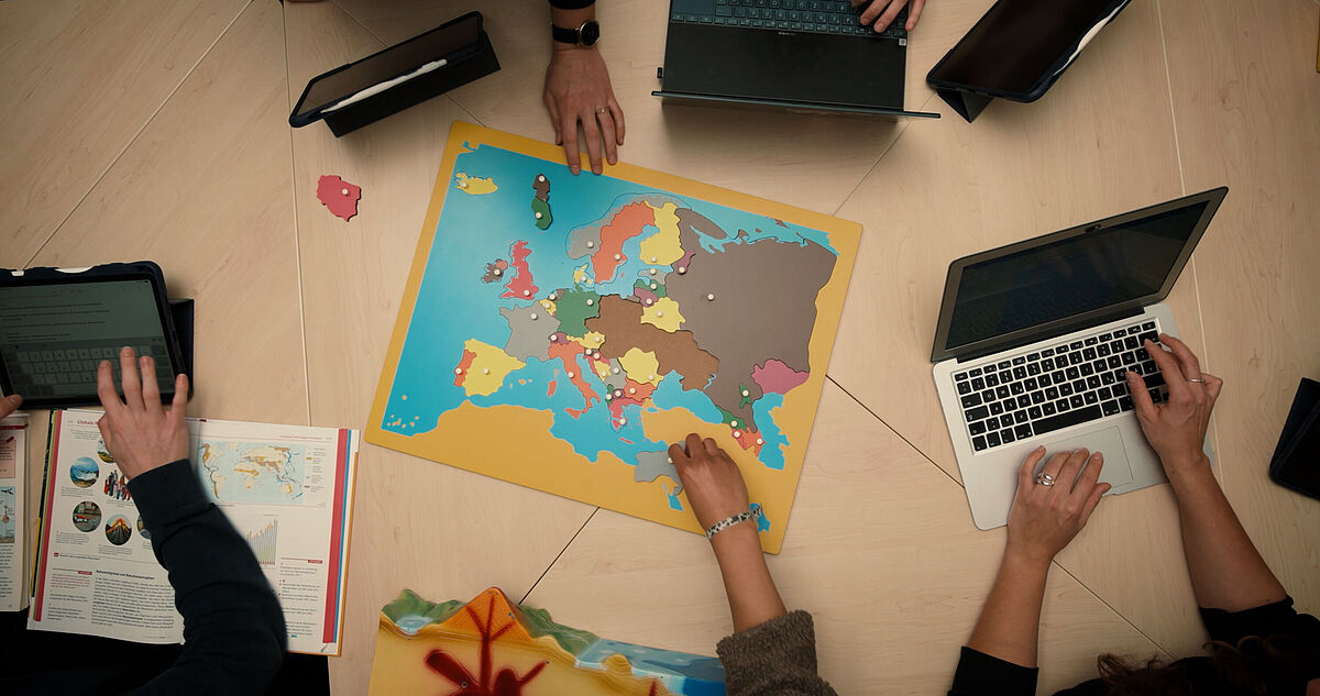 Geographie studieren Symbolbild Auf einem Tisch liegt eine Weltkarte mit diversten Pins. Darum sieht man die Hände von vier Menschen, die etwas auf ihrer Laptoptastatur tippen.