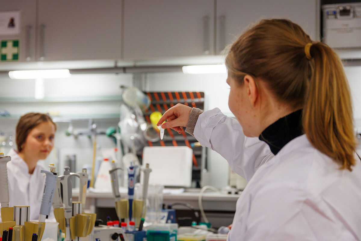 In eine Labor hält ein Mädchen im Laborkittel einn Glas mit einer flüssigen Substanz in ihrer Hand und betrachtet diese.