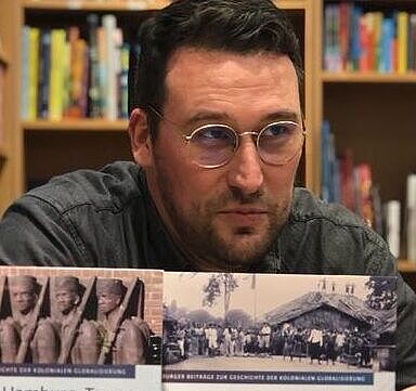 Dr. Kim Sebastian Todzi (Universität Hamburg) hält ein Buch in den Händen. Hinter ihm ist ein großes Bücherregal sichtbar.