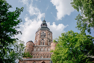 Blick auf den Turm des Domes St. Nikolai. Rechts und links stehen hohe Bäume mit grünem Laub..